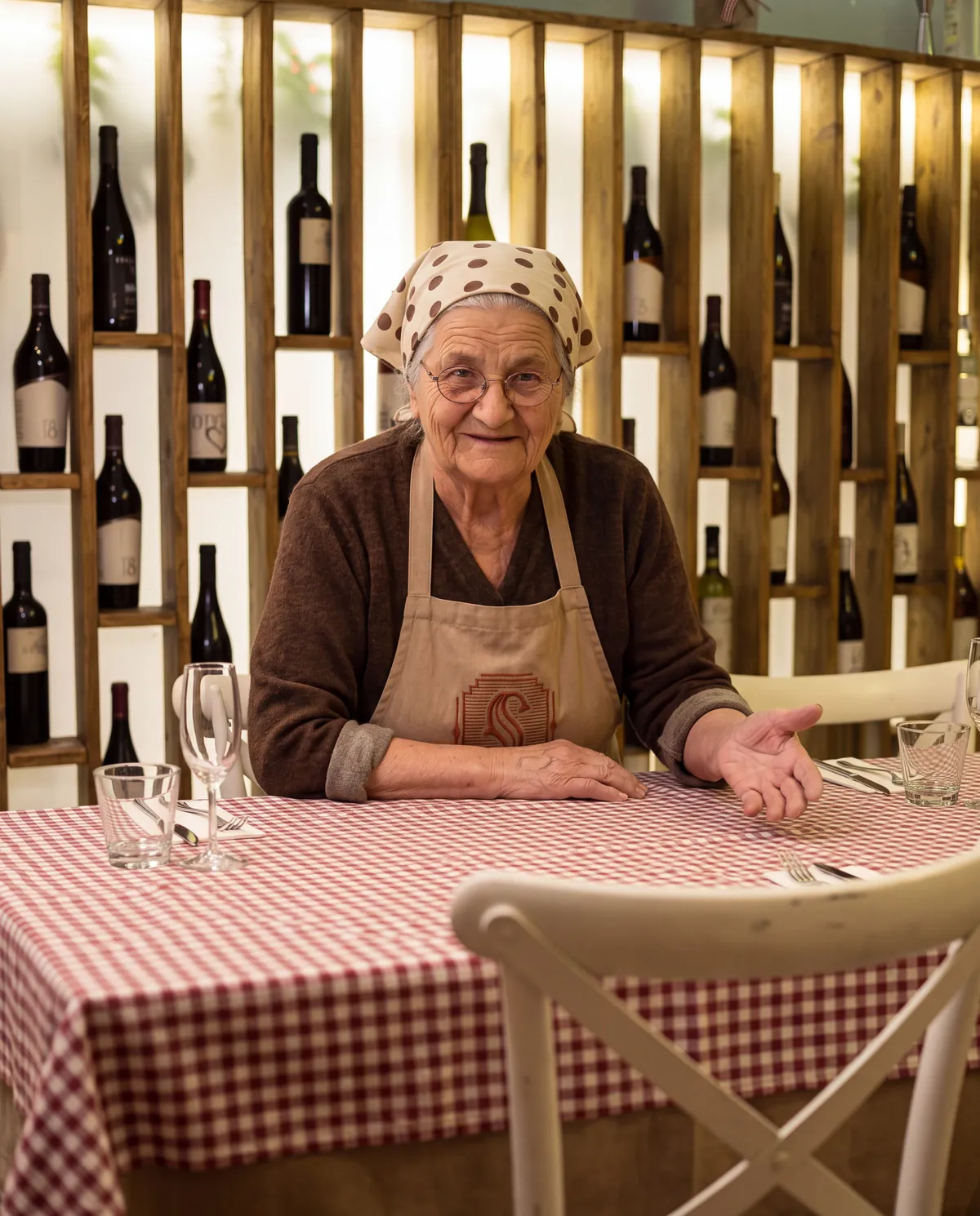 La nonna di aSud seduta a un tavolo con la tovaglia a quadri, davanti alla parete dei vini, che invita a sedersi
